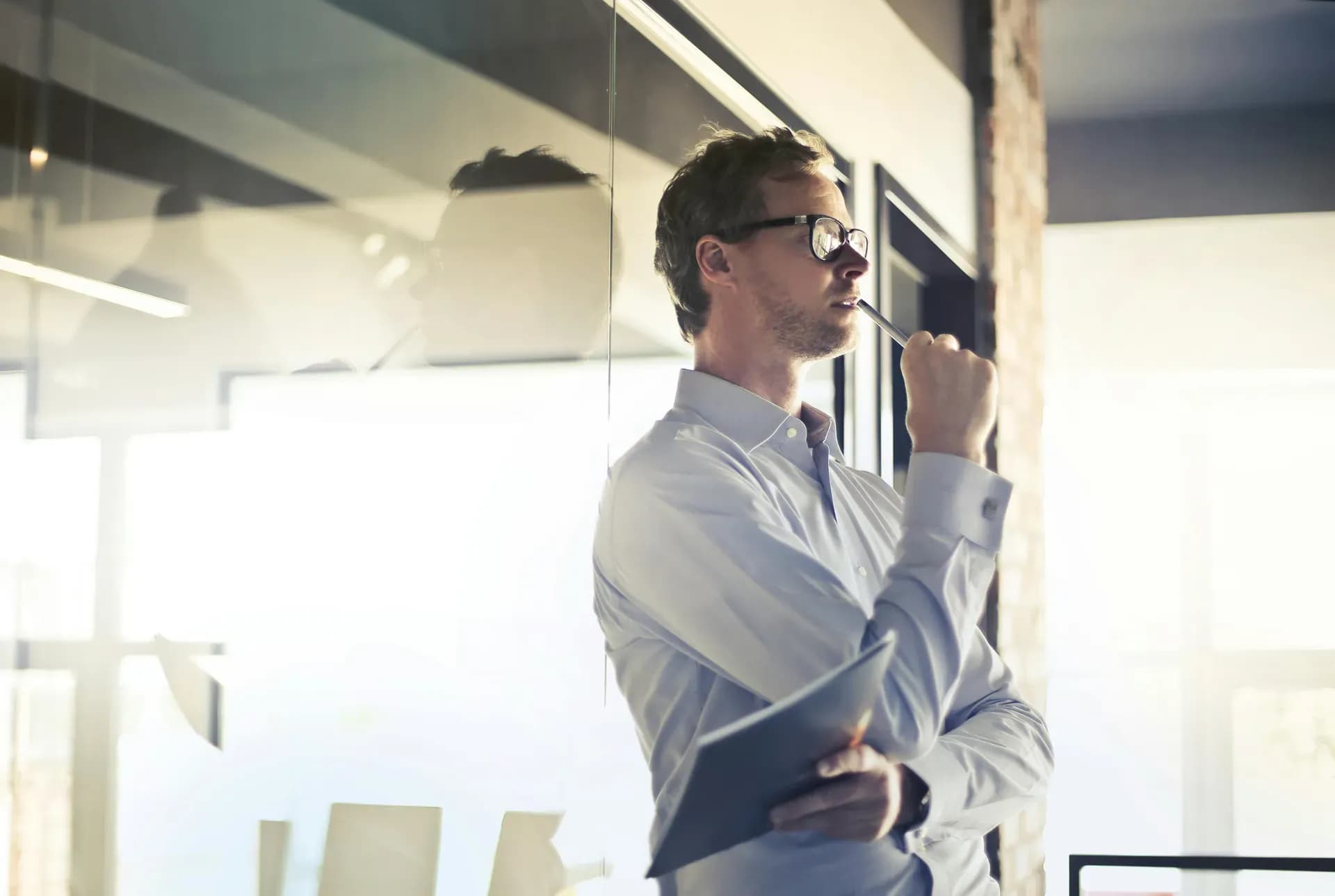 Thoughtful professional in glasses pausing at an office window, holding documents — reflecting on how their role has changed